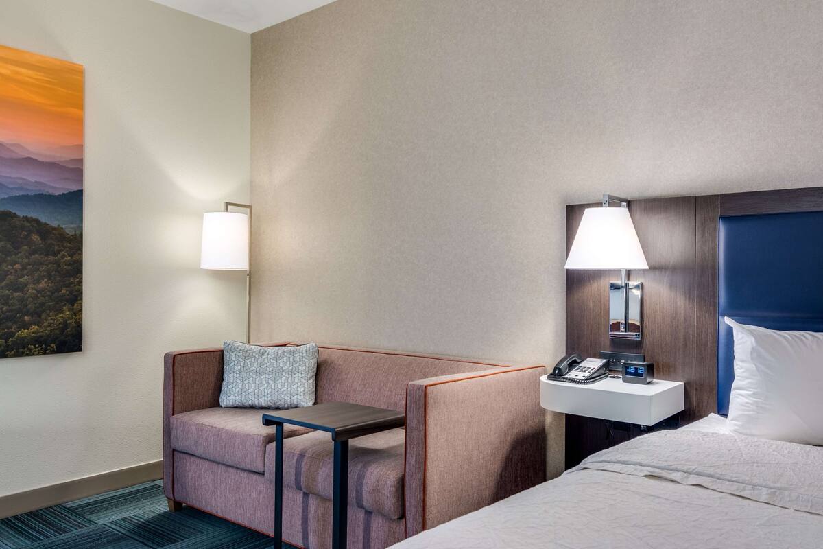 A hotel room with a bed, a desk with a lamp, a phone, and a painting of a sunset on the wall.