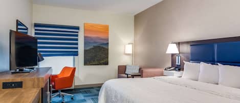 Habitación de hotel con cama, escritorio con televisor, silla, mesita de noche y un cuadro en la pared.