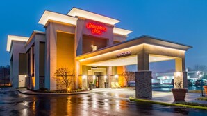 Ett modernt Hampton Inn-hotell med en väl upplyst entré, en tydligt markerad parkeringsplats och en klar himmel.