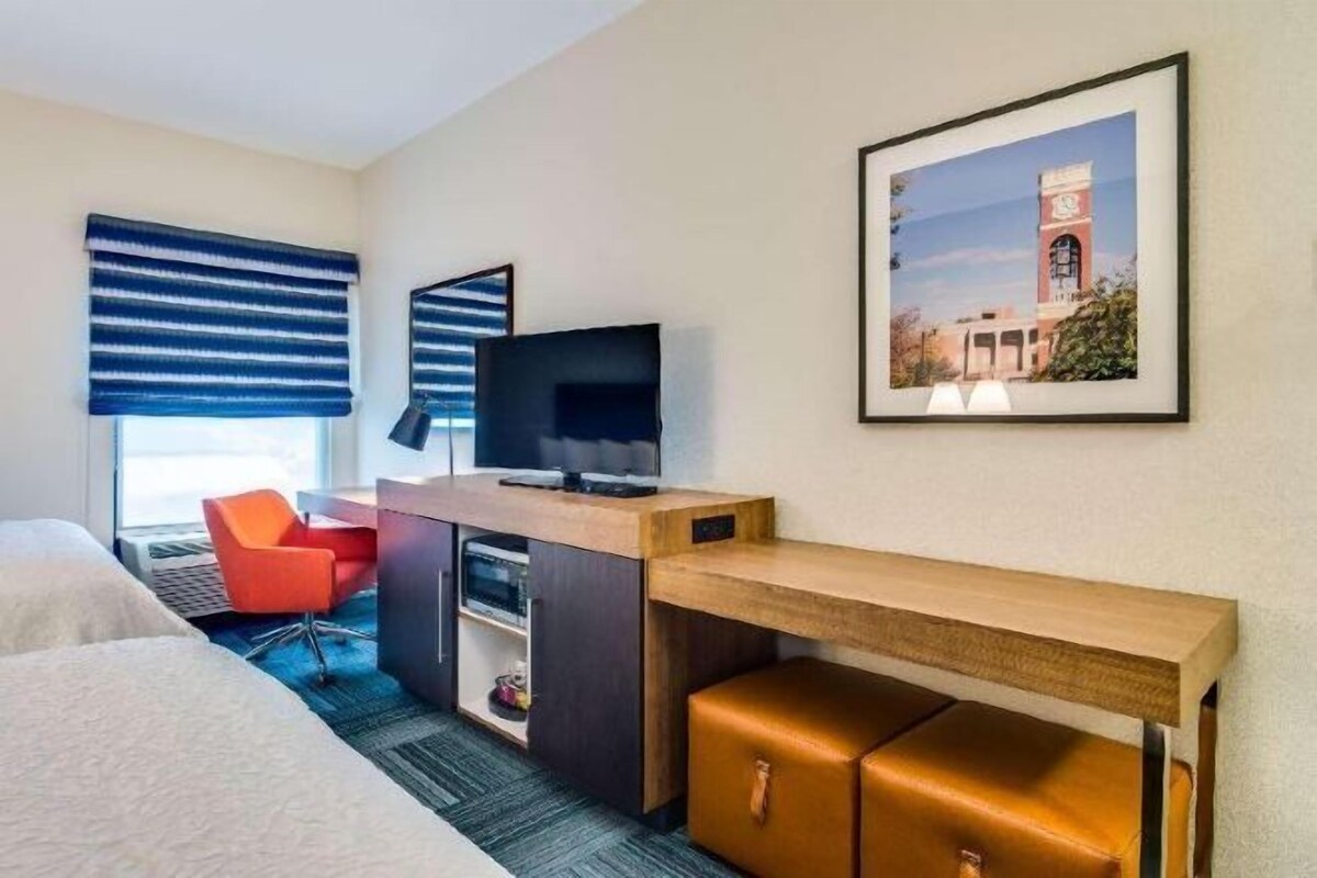 Quarto de hotel com cama, escrivaninha com televisão, cadeira, mesa pequena e um quadro na parede.