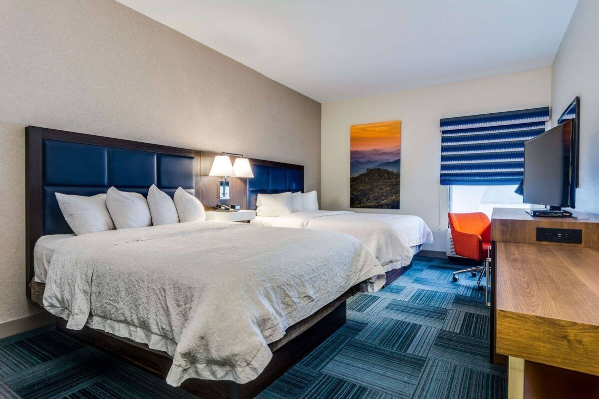 A hotel room with two beds, a desk, a TV, and a painting on the wall.