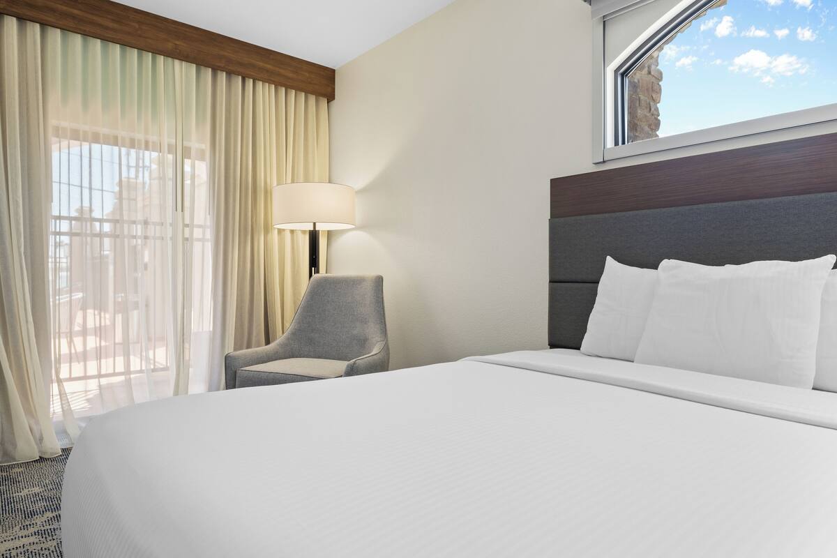 A hotel room with a large bed, a chair, a lamp, and a window with a view of the sky.