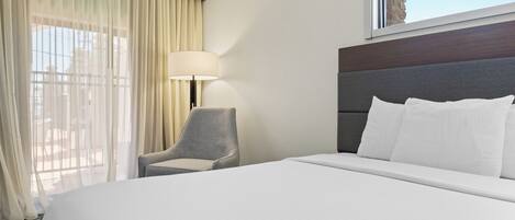 Habitación de hotel con una cama grande, una silla, una lámpara y un ventanal con vistas al cielo.
