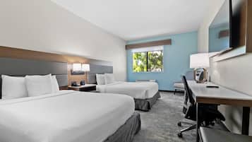 A hotel room with two beds, a desk, a chair, a TV, and a window with a view of trees.