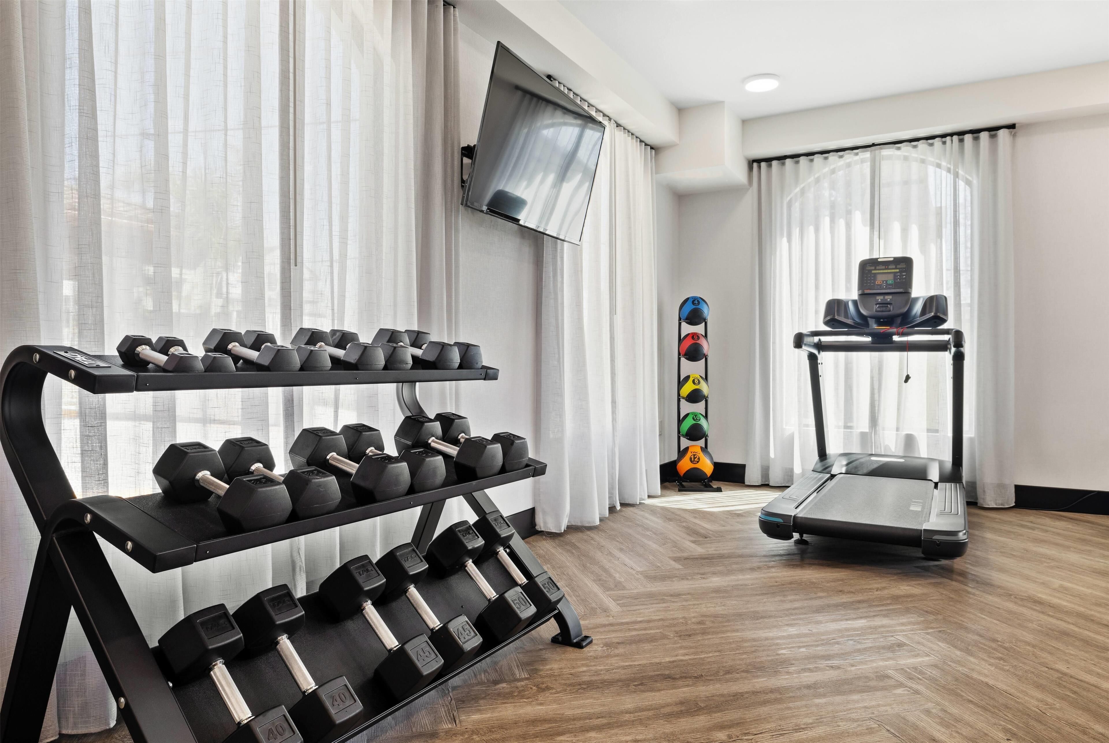 Un gimnasio con una cinta de correr, mancuernas y un televisor en la pared.