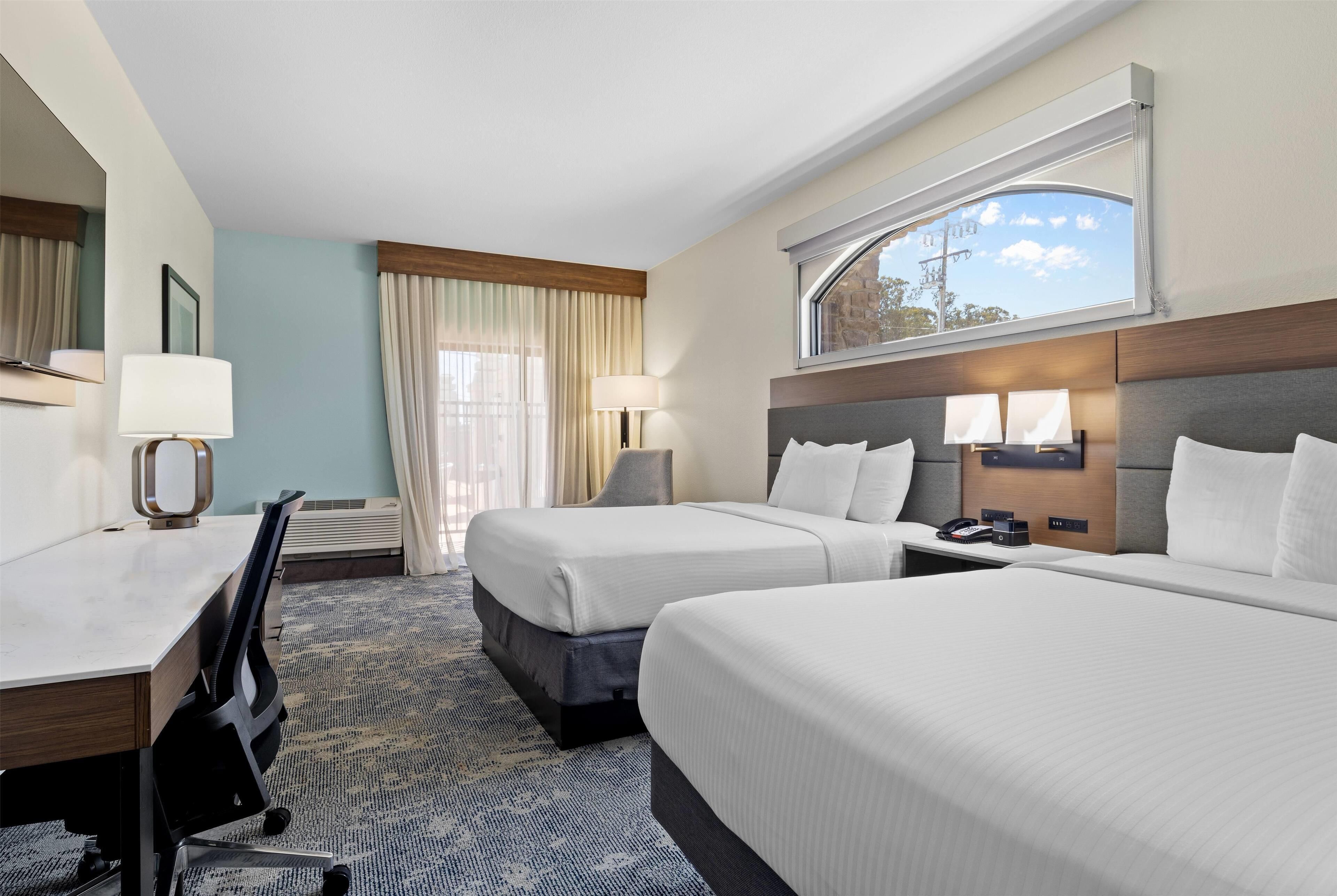 A hotel room with two beds, a desk, a chair, a lamp, and a window with curtains.