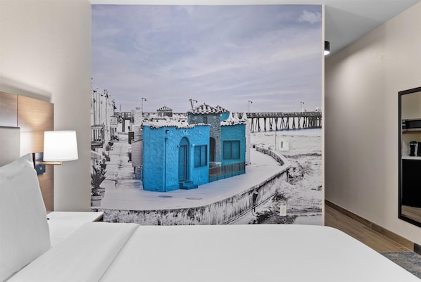 Habitación de hotel con un amplio mural en la pared que muestra una escena costera, una cama, una mesita de noche y una lámpara.
