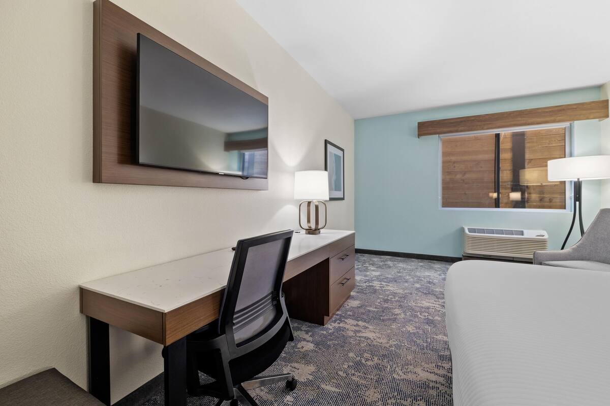 A hotel room with a desk, chair, bed, TV, and a window with blinds.