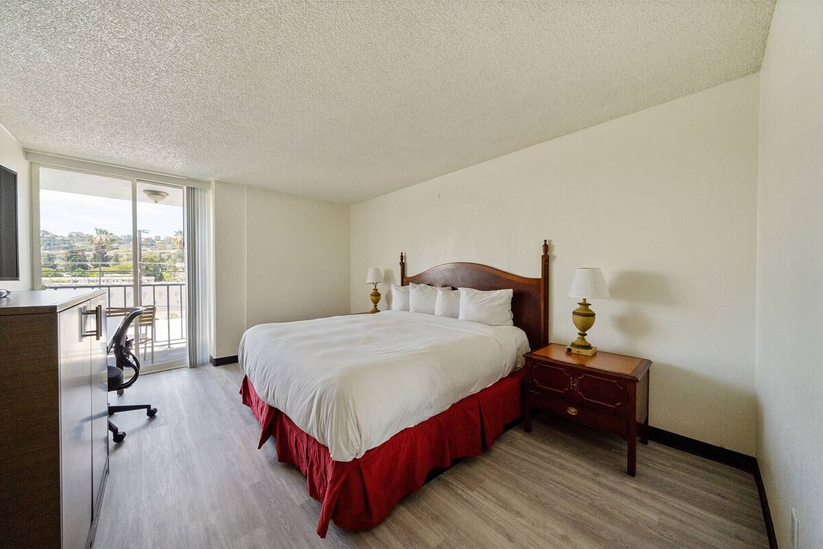 A hotel room with a large bed, a desk, and a chair. There is a window with a view of the outdoors.