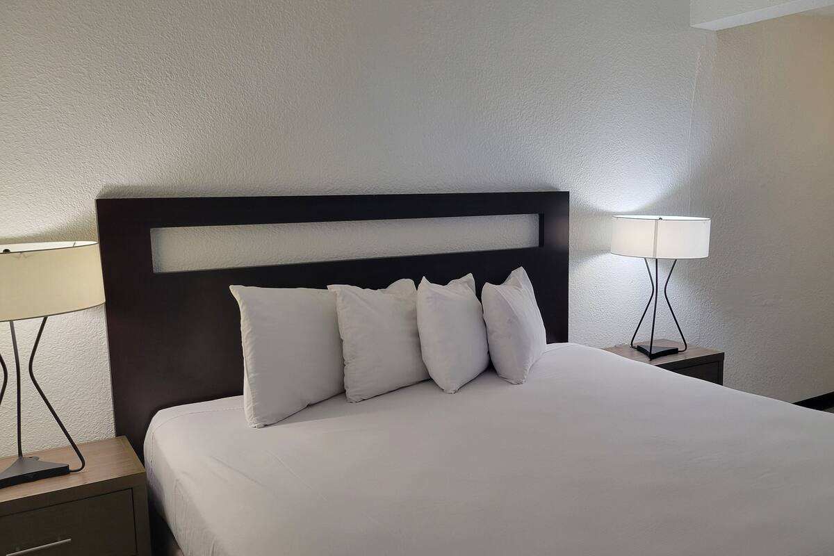 A neatly made bed with white linens and a dark headboard. There are four pillows on the bed. Two bedside tables flank the bed, each with a lamp. The room has a simple, clean design with white walls and a wooden floor.