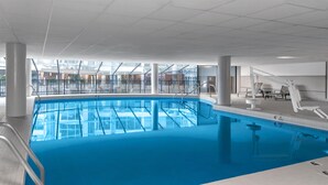 An indoor swimming pool with clear blue water, surrounded by white tiled walls and a ceiling with a grid pattern.