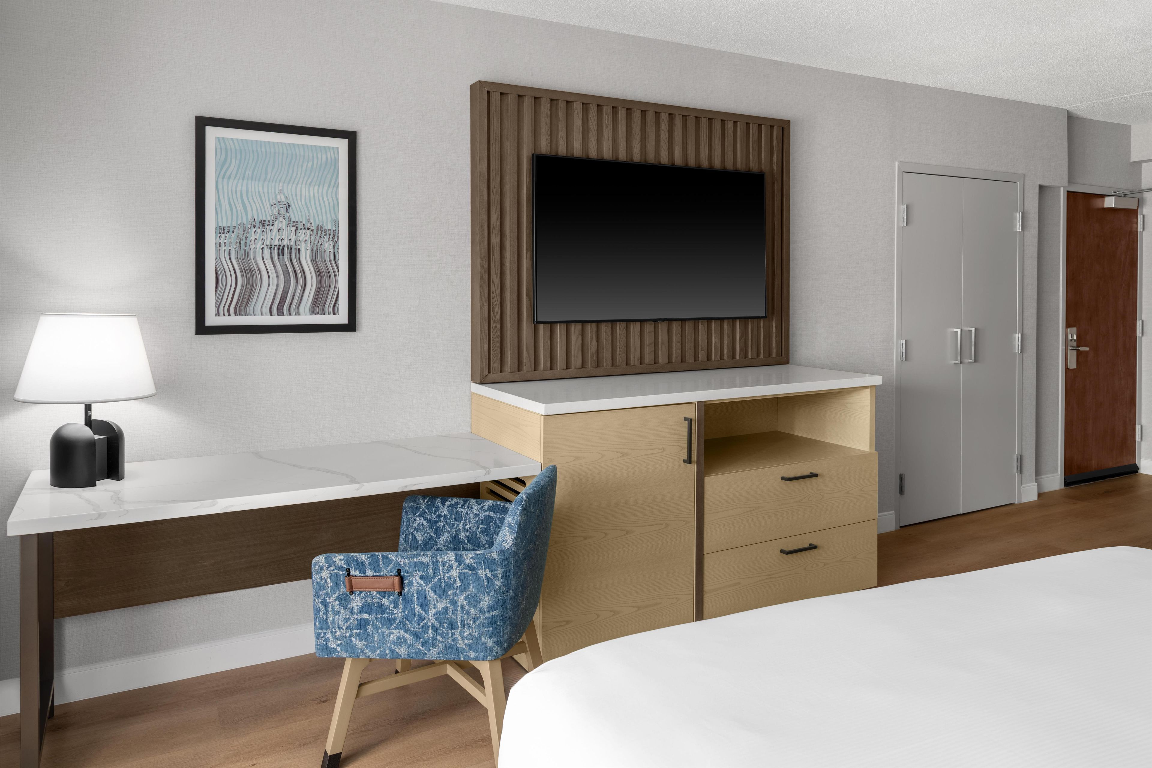 A hotel room with a bed, a desk with a lamp, a chair, a wardrobe, and a television on the wall.