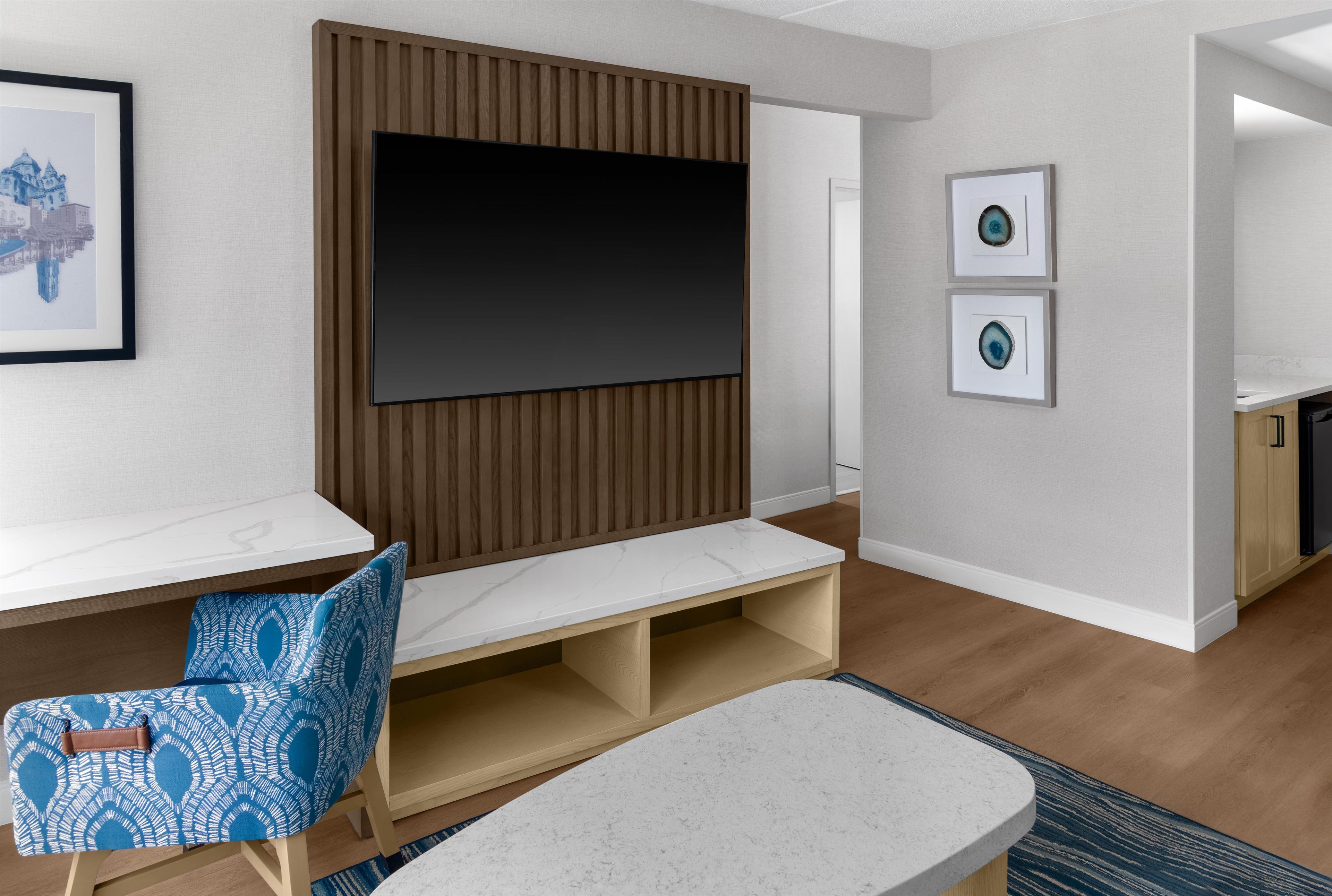 A modern living room with a flat-screen TV, a wooden panel wall, a white bench, a blue patterned chair, and two framed artworks.