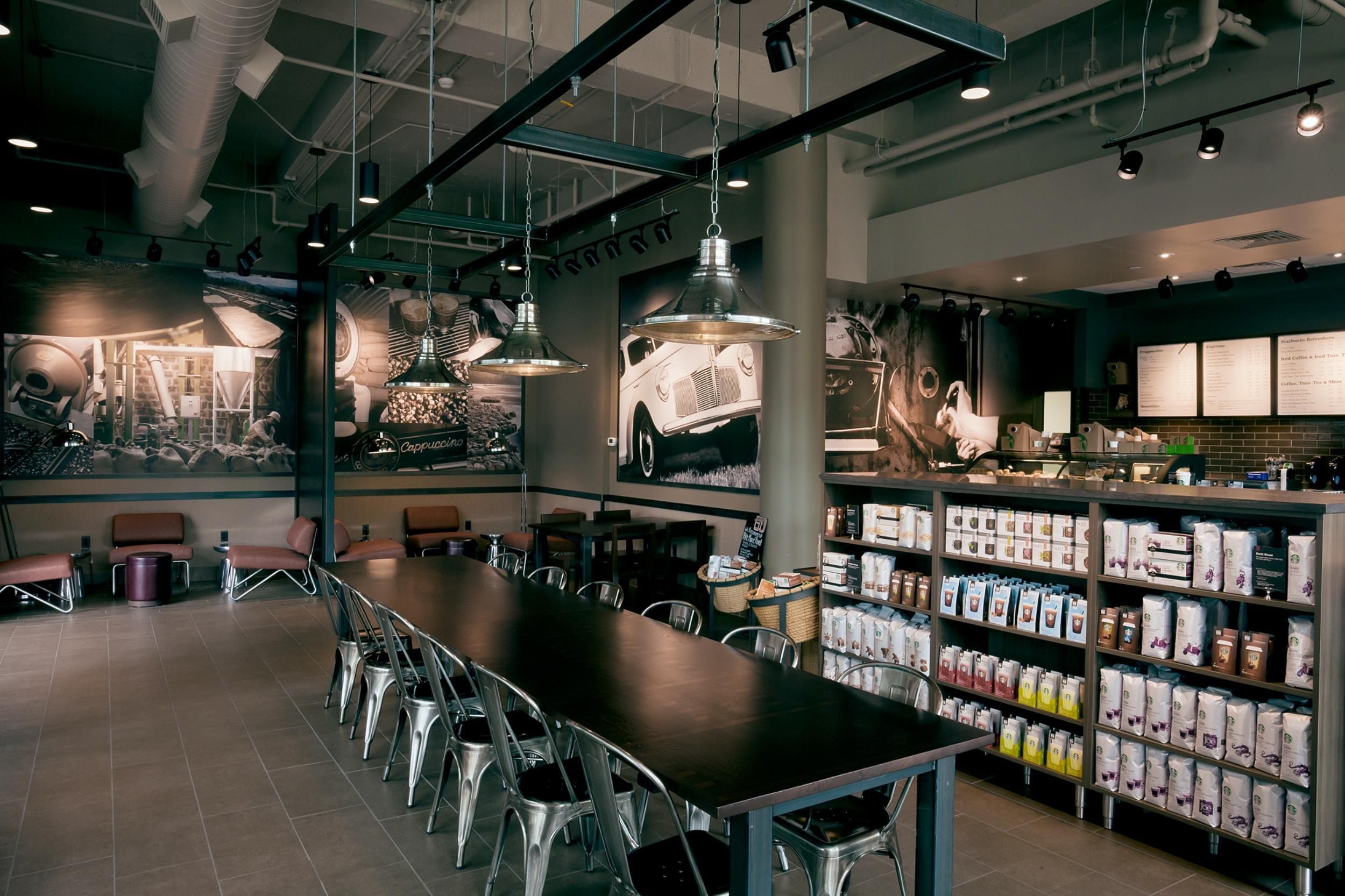 Un interno di caffetteria moderna con un lungo tavolo condiviso, sedie in metallo, lampade a sospensione e un'esposizione di opere d'arte a tema caffè.