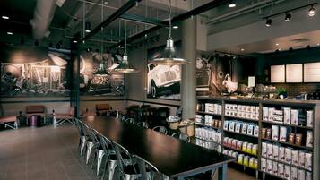 Un interno di caffetteria moderna con un lungo tavolo condiviso, sedie in metallo, lampade a sospensione e un'esposizione di opere d'arte a tema caffè.