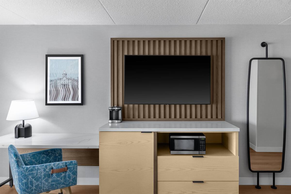 Ein modernes Hotelzimmer mit einem großen Flachbildfernseher an einer Wand aus Holzpaneelen, einem kleinen Schreibtisch mit Lampe, einer Mikrowelle und einem blauen Stuhl.