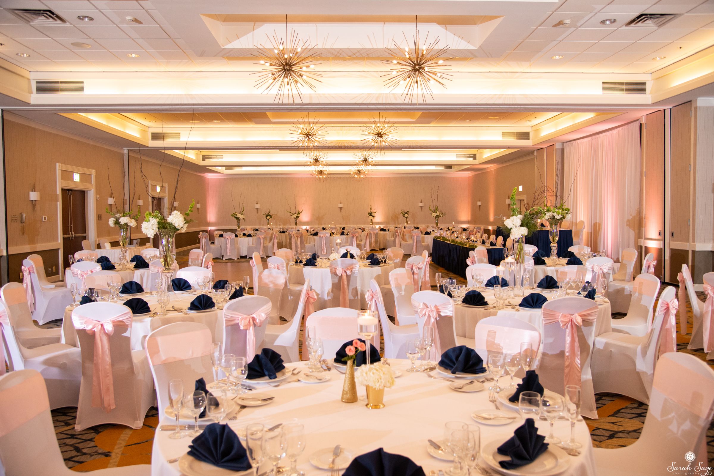 Une salle de banquet avec des tables rondes, des chaises blanches et bleu marine, et des centres de table floraux.