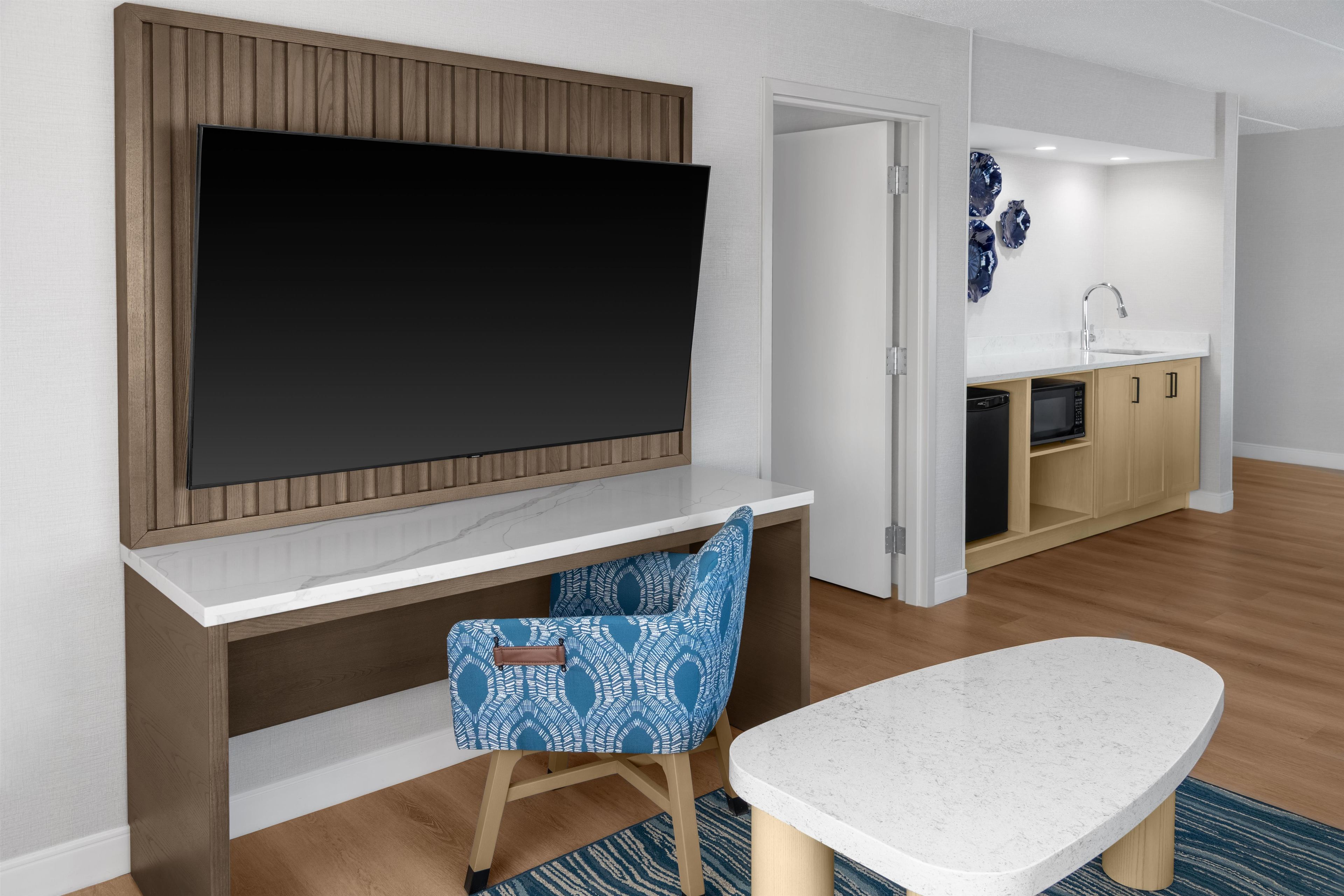 Una sala moderna con un televisor de pantalla plana montado en una pared de paneles de madera, una mesa de comedor con cubierta de mármol blanco y una silla azul estampada.