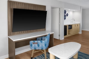 A modern living room with a flat-screen TV mounted on a wooden panel wall, a dining table with a white marble top, and a blue patterned chair. - Doubletree Hotel South Bend (South Bend)