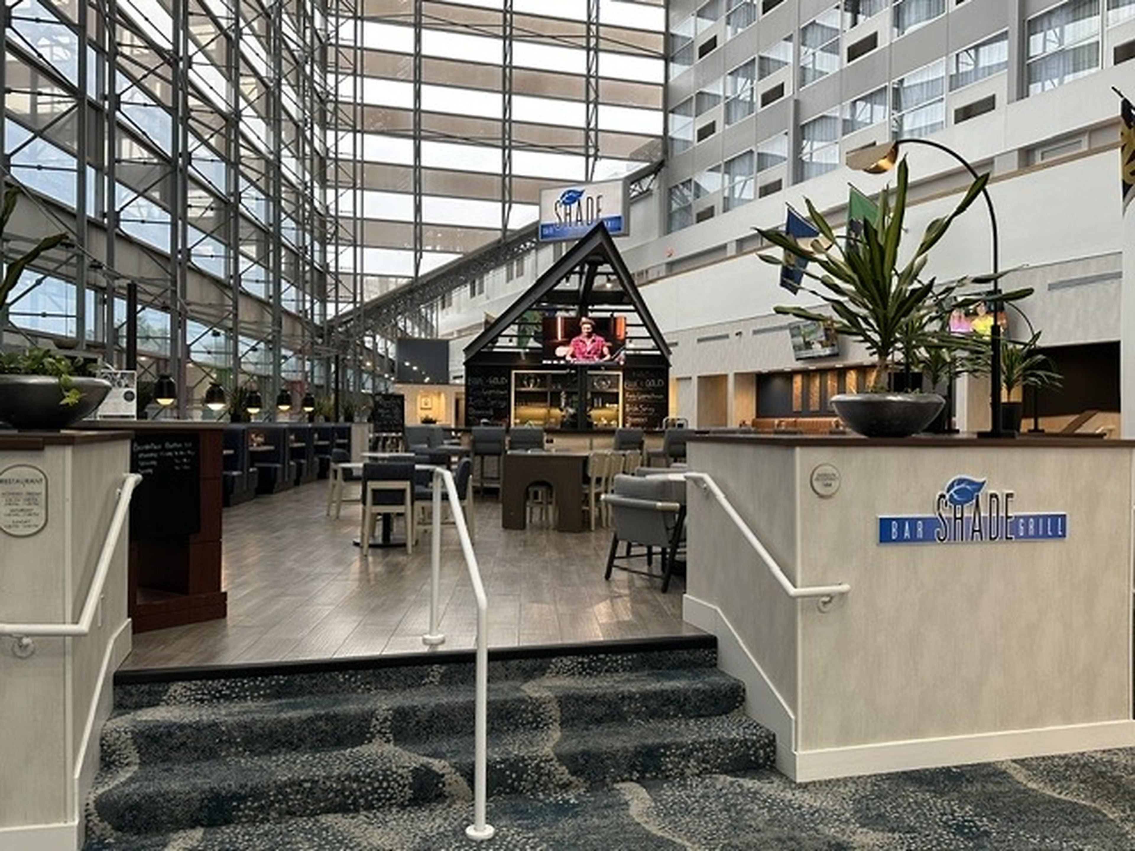 A modern hotel lobby with a bar and seating area, a staircase leading to upper floors, and a large glass atrium.