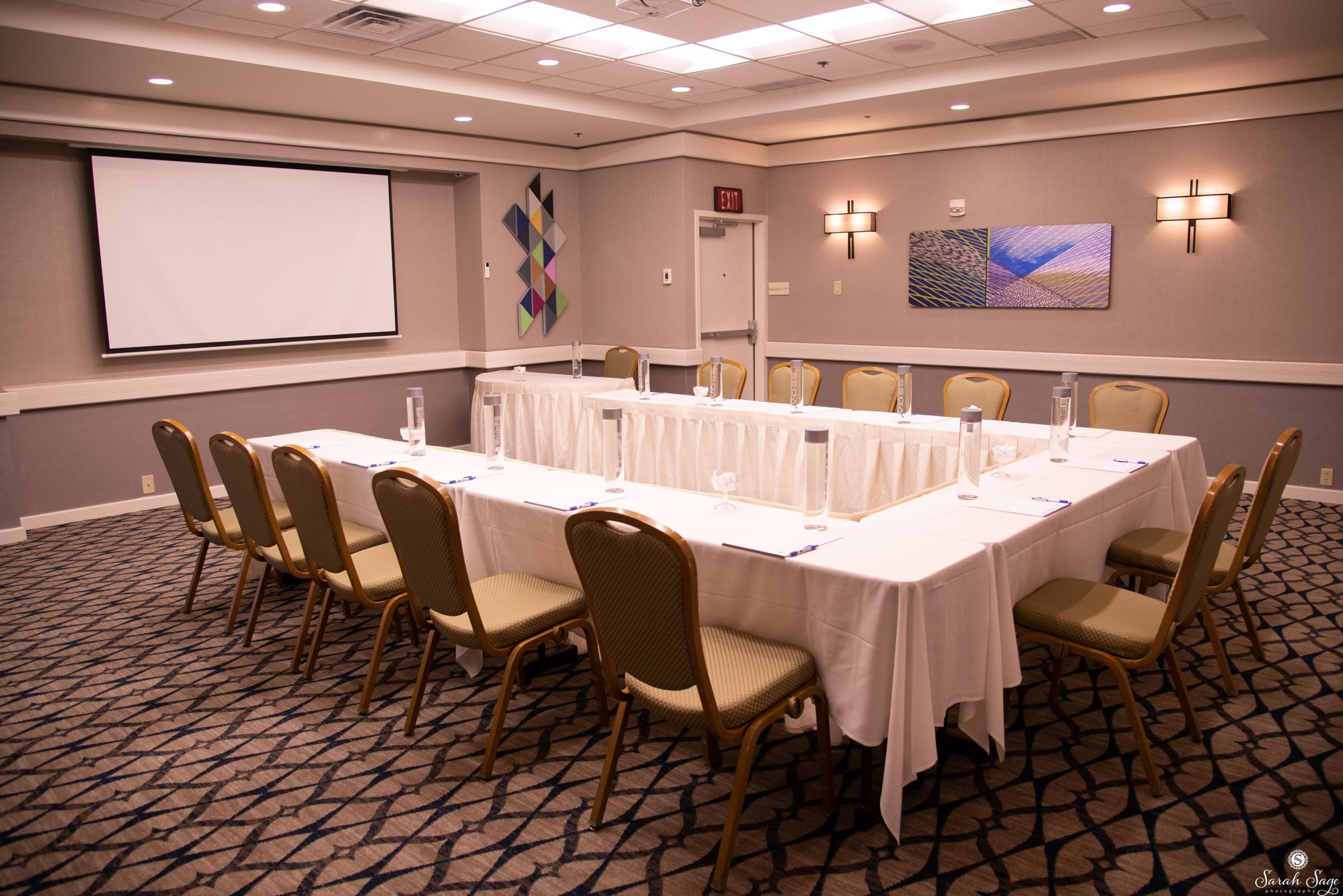 A conference room with a long table, chairs, a projector screen, and wall art.