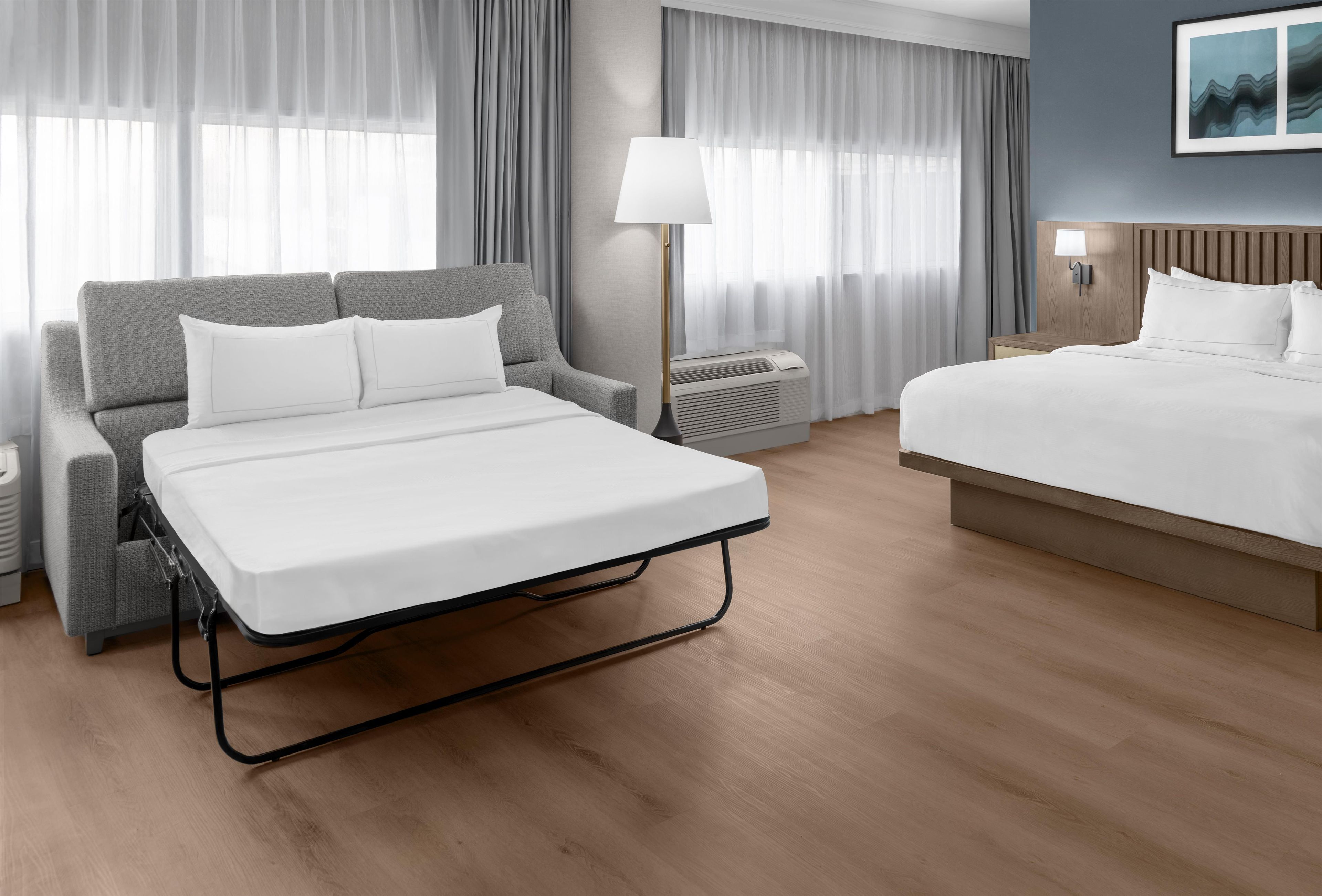 A hotel room with a bed, a sofa, a lamp, and a wall-mounted air conditioner.