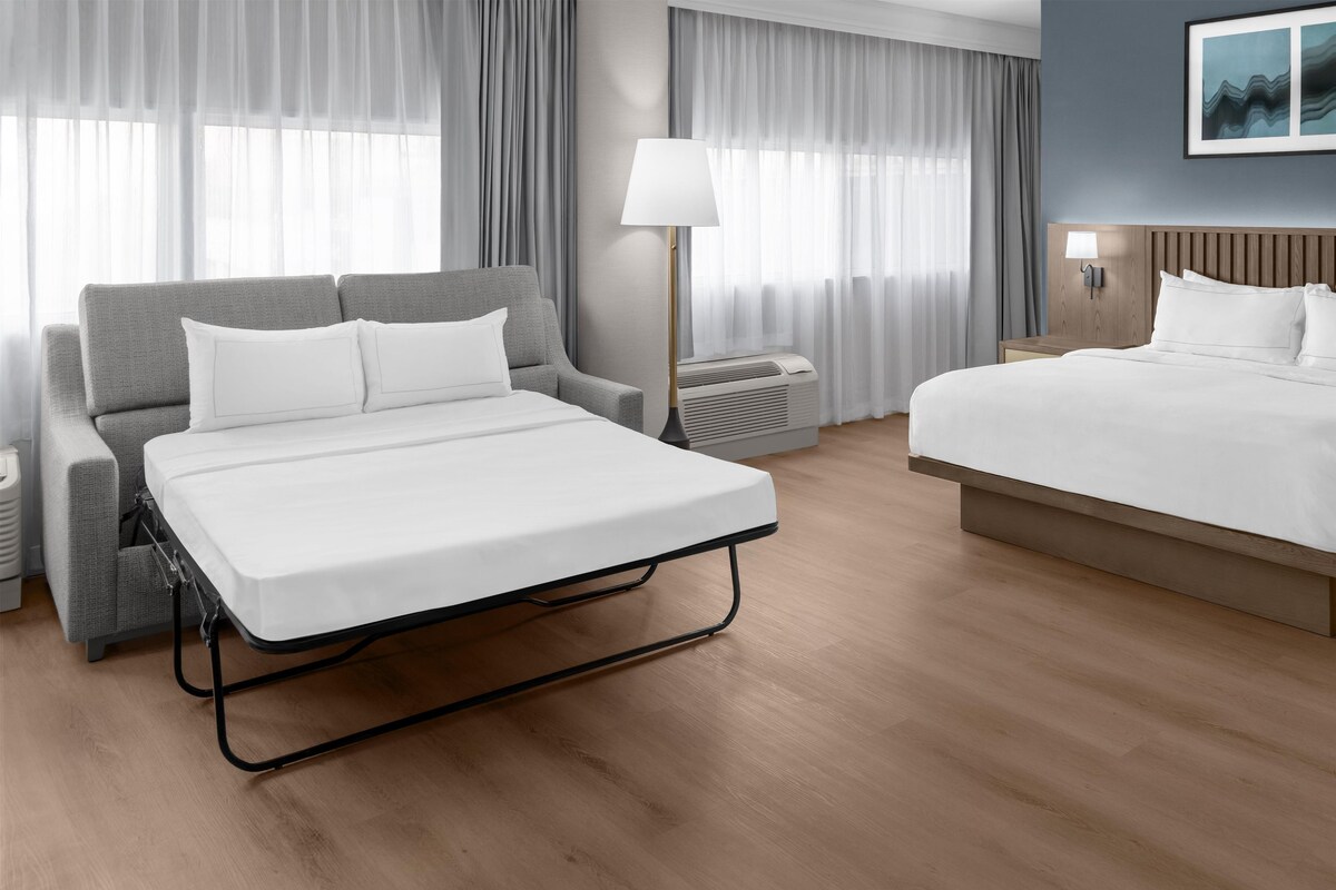 A hotel room with a bed, a sofa, a lamp, and a wall-mounted air conditioner.
