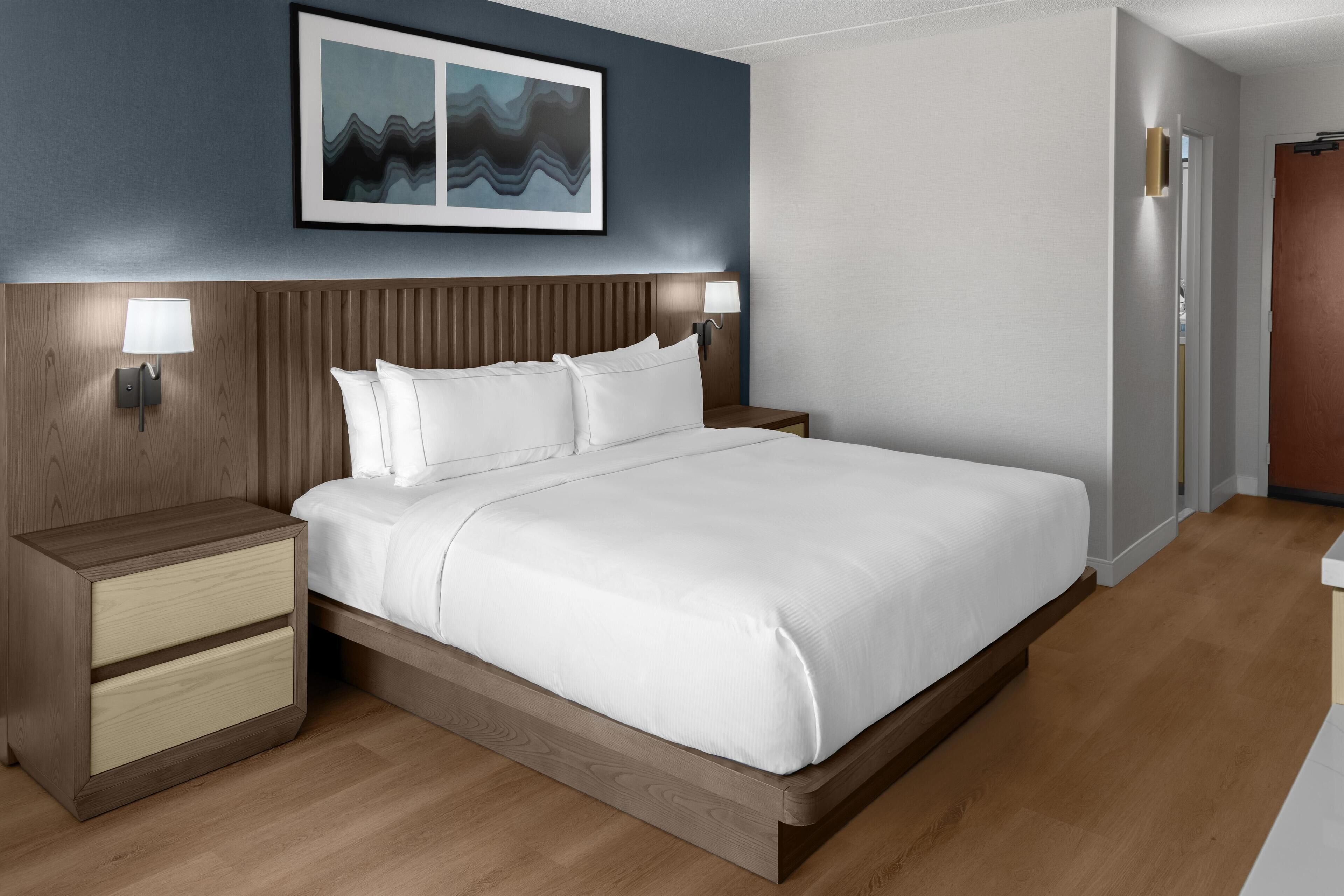 Une chambre d’hôtel moderne avec une tête de lit en bois, un grand lit avec du linge de lit blanc, deux tables de chevet avec des lampes et une œuvre d’art encadrée au-dessus du lit.