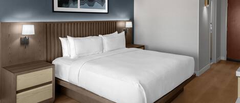 A modern hotel room with a wooden headboard, a large bed with white linens, two bedside tables with lamps, and a framed artwork above the bed.
