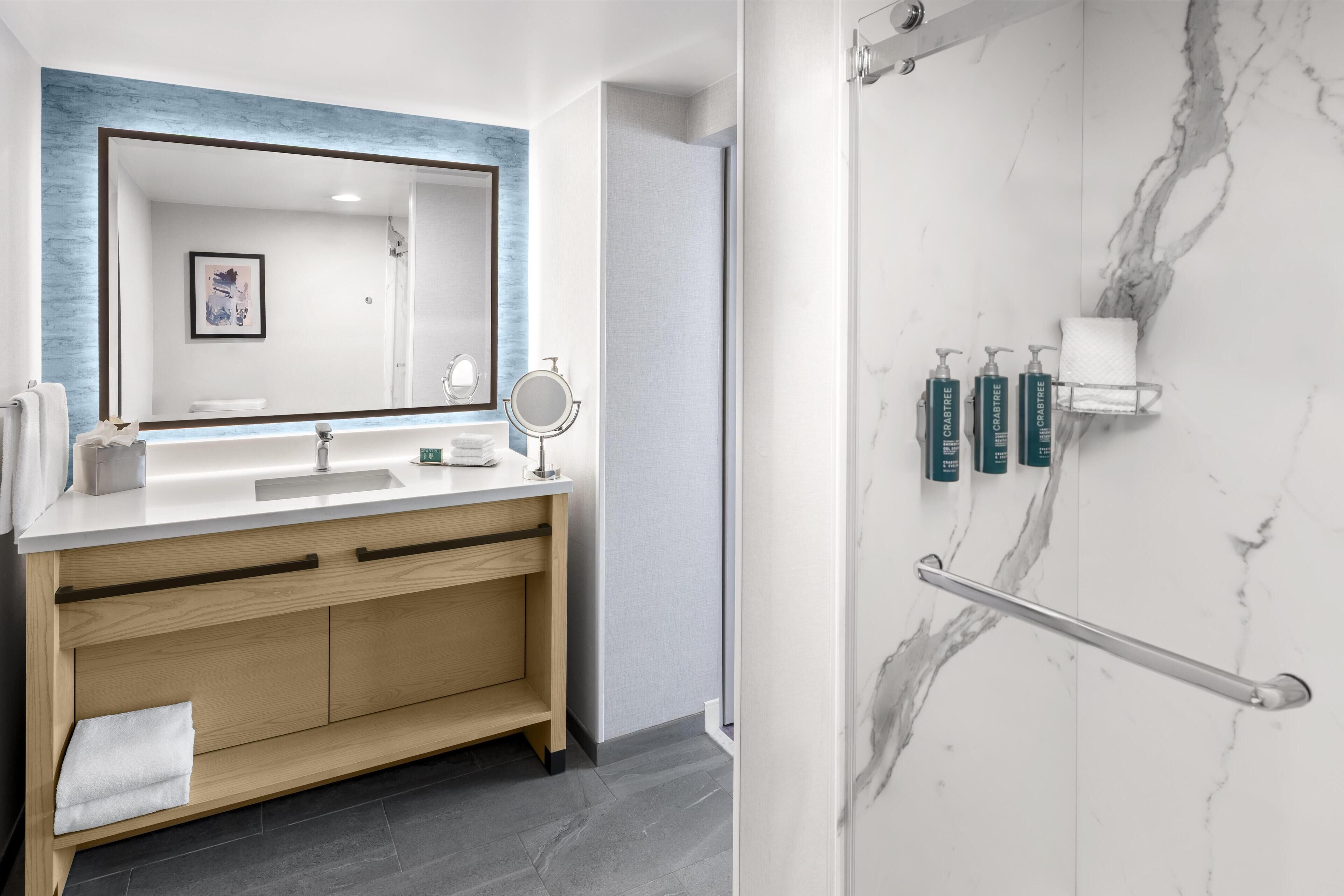 A modern bathroom with a large mirror, wooden vanity, and a marble shower area.