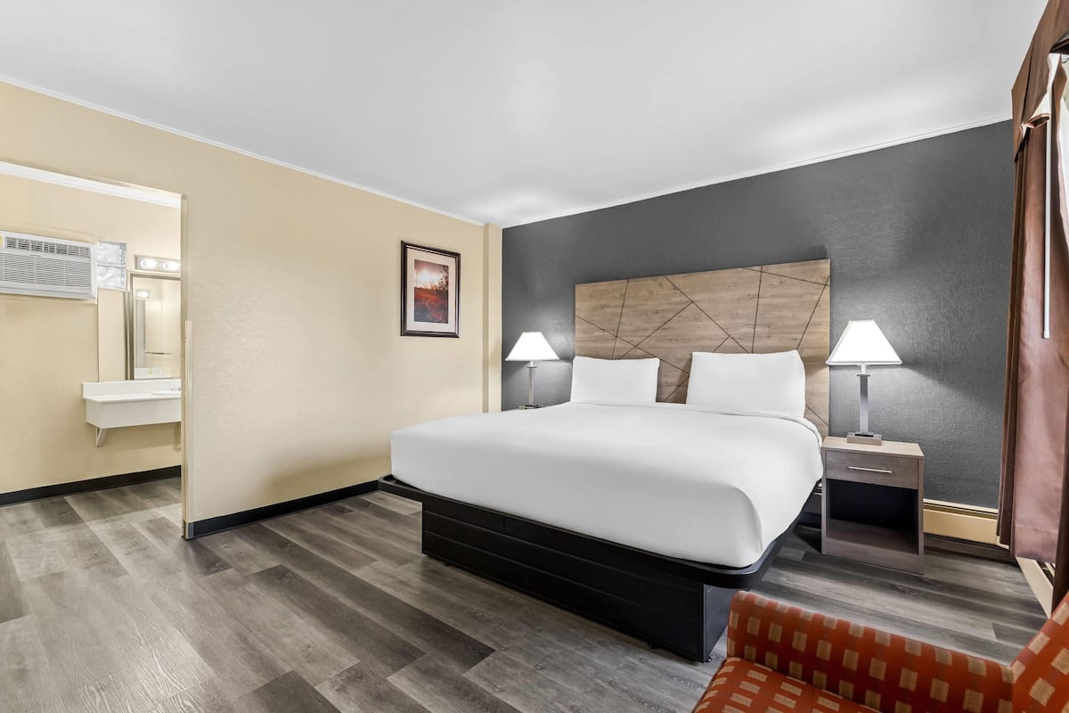 Ein Hotelzimmer mit einem großen Bett, Nachttischlampen, einem Nachttisch, einem Sessel, einem Spiegel und einer Wandklimaanlage.