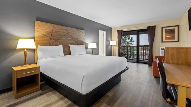 A hotel room with a large bed, two bedside tables with lamps, a wooden headboard, a balcony with a view, and a desk with a chair. - Travelodge by Wyndham Rapid City (Rapid City)