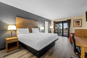 A hotel room with a large bed, two bedside tables with lamps, a wooden headboard, a balcony with a view, and a desk with a chair. - Travelodge by Wyndham Rapid City (Rapid City)