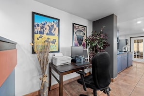 A home office with a desk, computer, and printer. There are two framed posters on the wall, a vase with dried plants, and a kitchen area in the background. - Travelodge by Wyndham Rapid City (Rapid City)