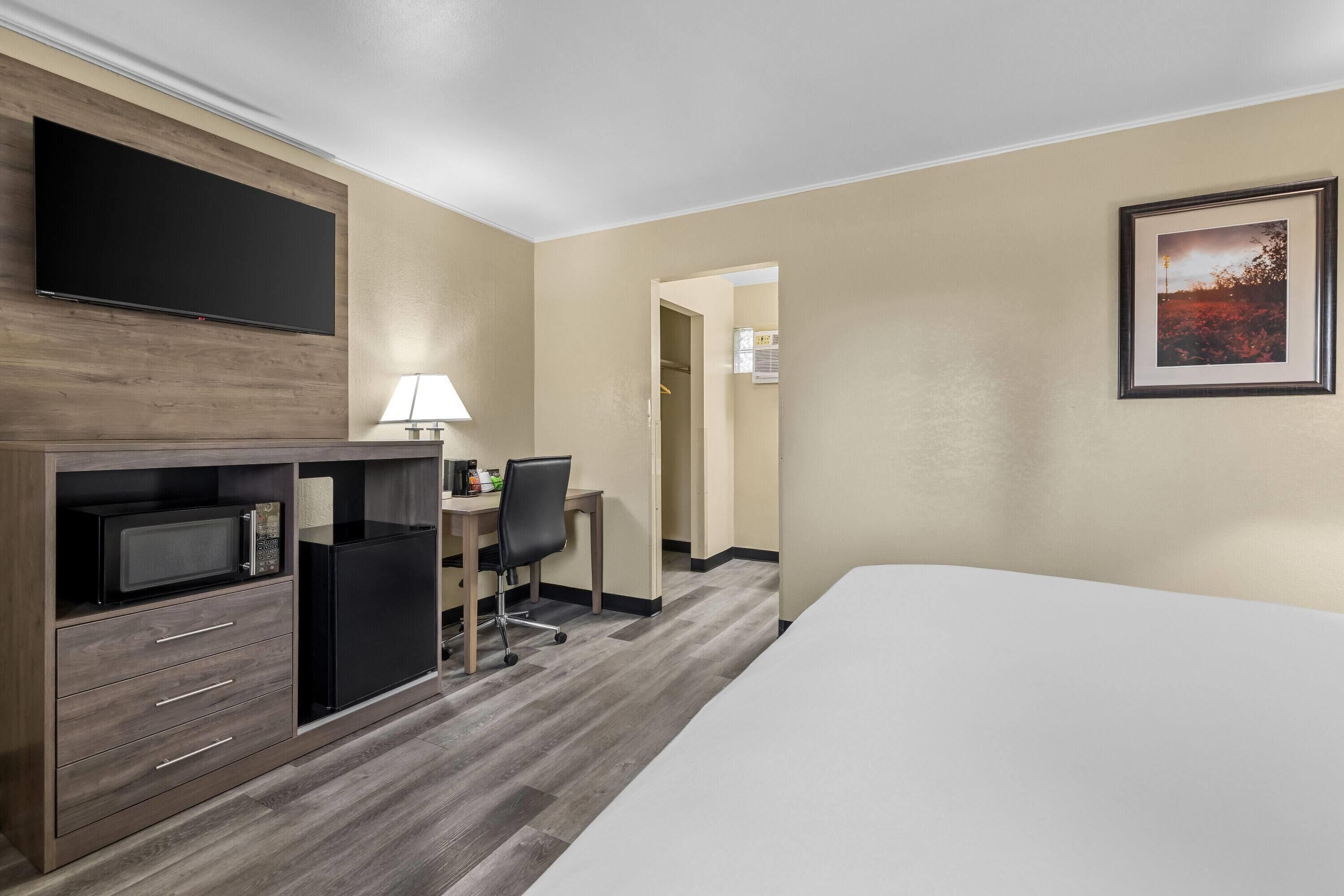 A hotel room with a bed, a desk with a chair, a microwave, a refrigerator, and a flat-screen TV mounted on the wall.