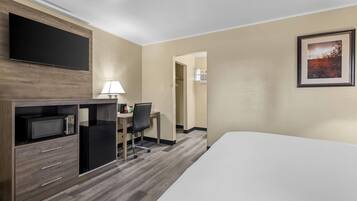 A hotel room with a bed, a desk with a chair, a microwave, a refrigerator, and a flat-screen TV mounted on the wall.