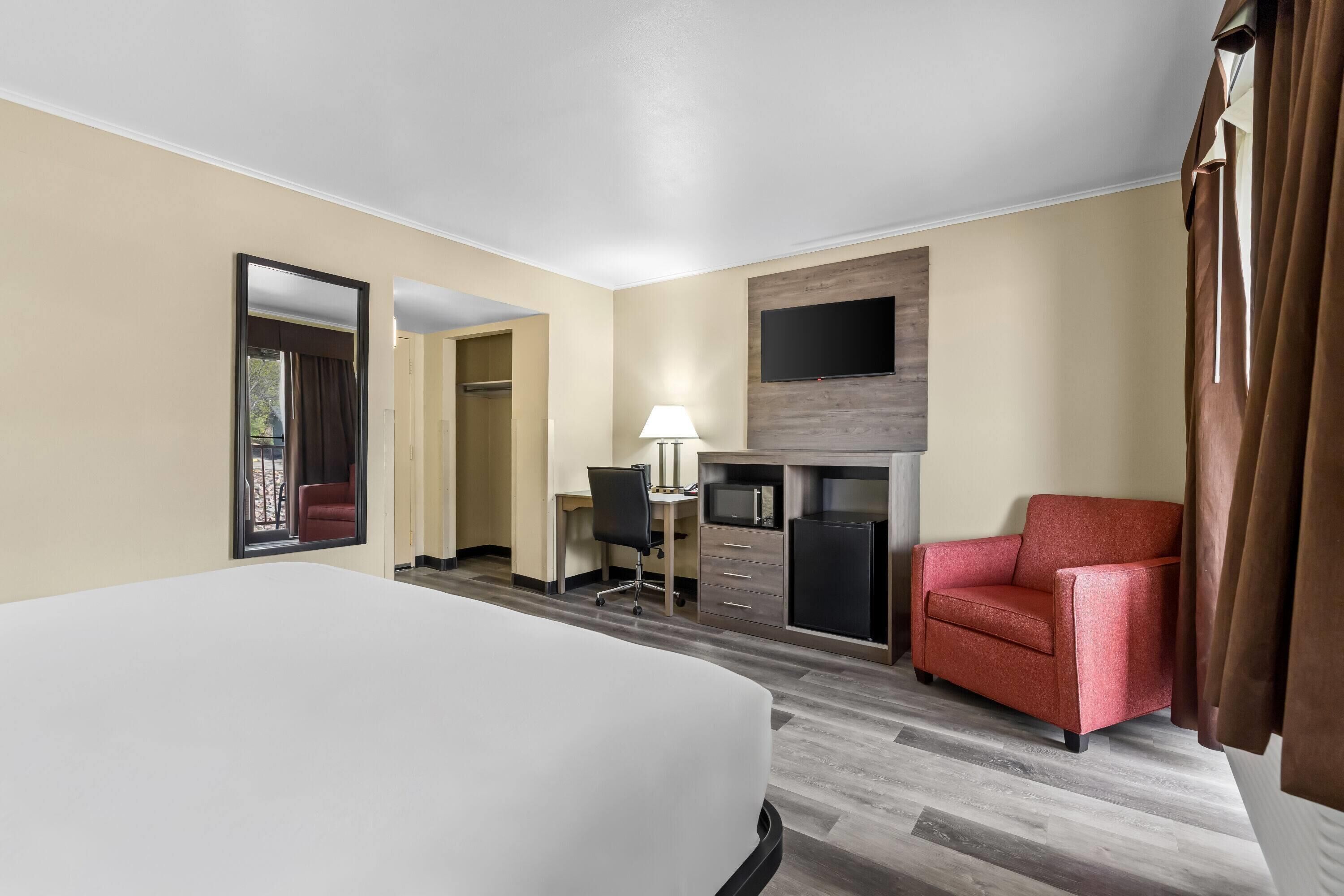 A hotel room with a bed, a desk with a chair, a TV mounted on the wall, and a red armchair.