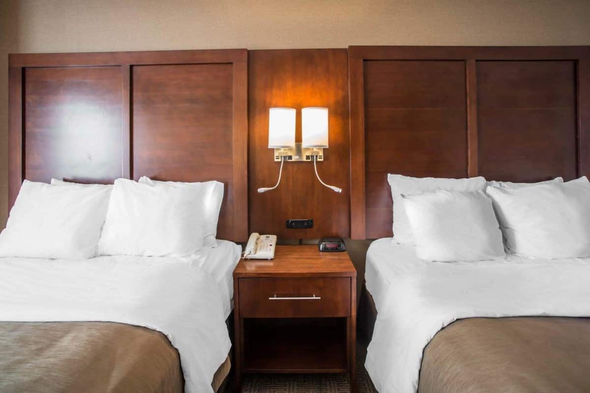 A hotel room with two beds, a nightstand with a phone and a lamp, and a wall-mounted light fixture.