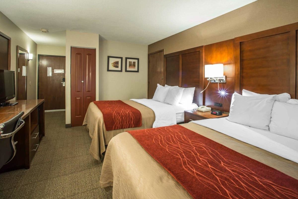 A hotel room with two beds, a desk with a television, a bedside table with a lamp, and a door.