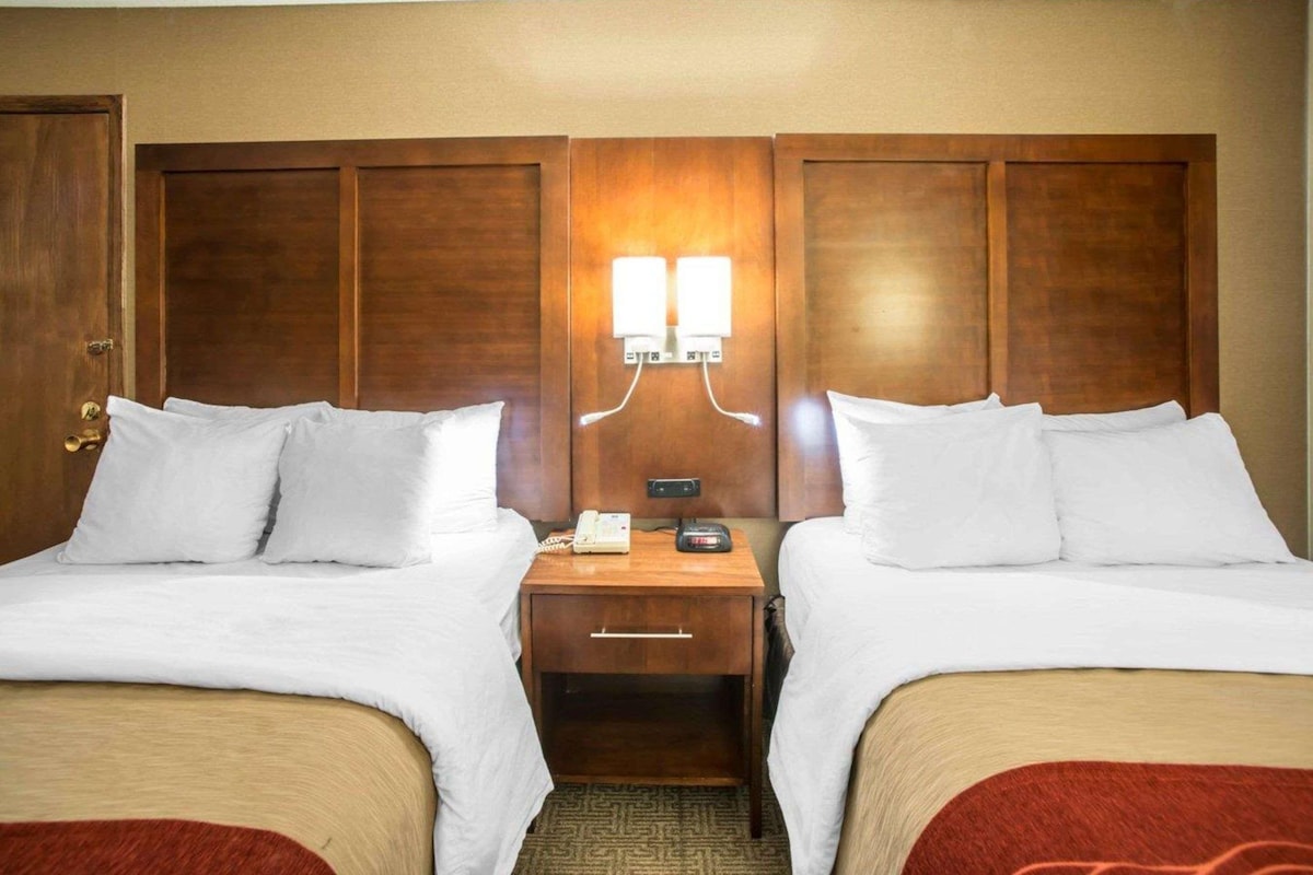 A hotel room with two beds, a nightstand with a phone and a lamp, and a wooden headboard.
