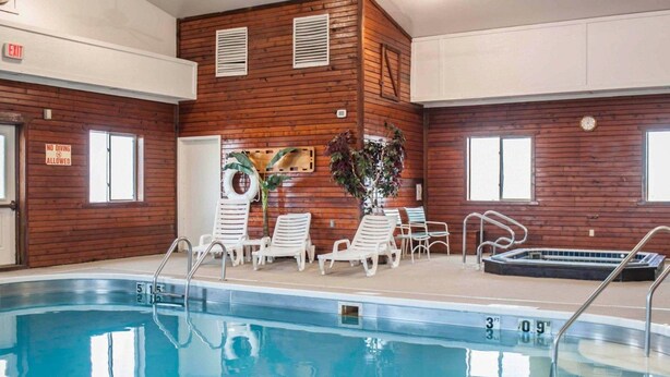 An indoor swimming pool with lounge chairs and a hot tub, surrounded by wooden walls and windows.