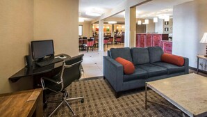 A hotel room with a desk, computer, and a sofa. There is a dining area in the background with chairs and tables.