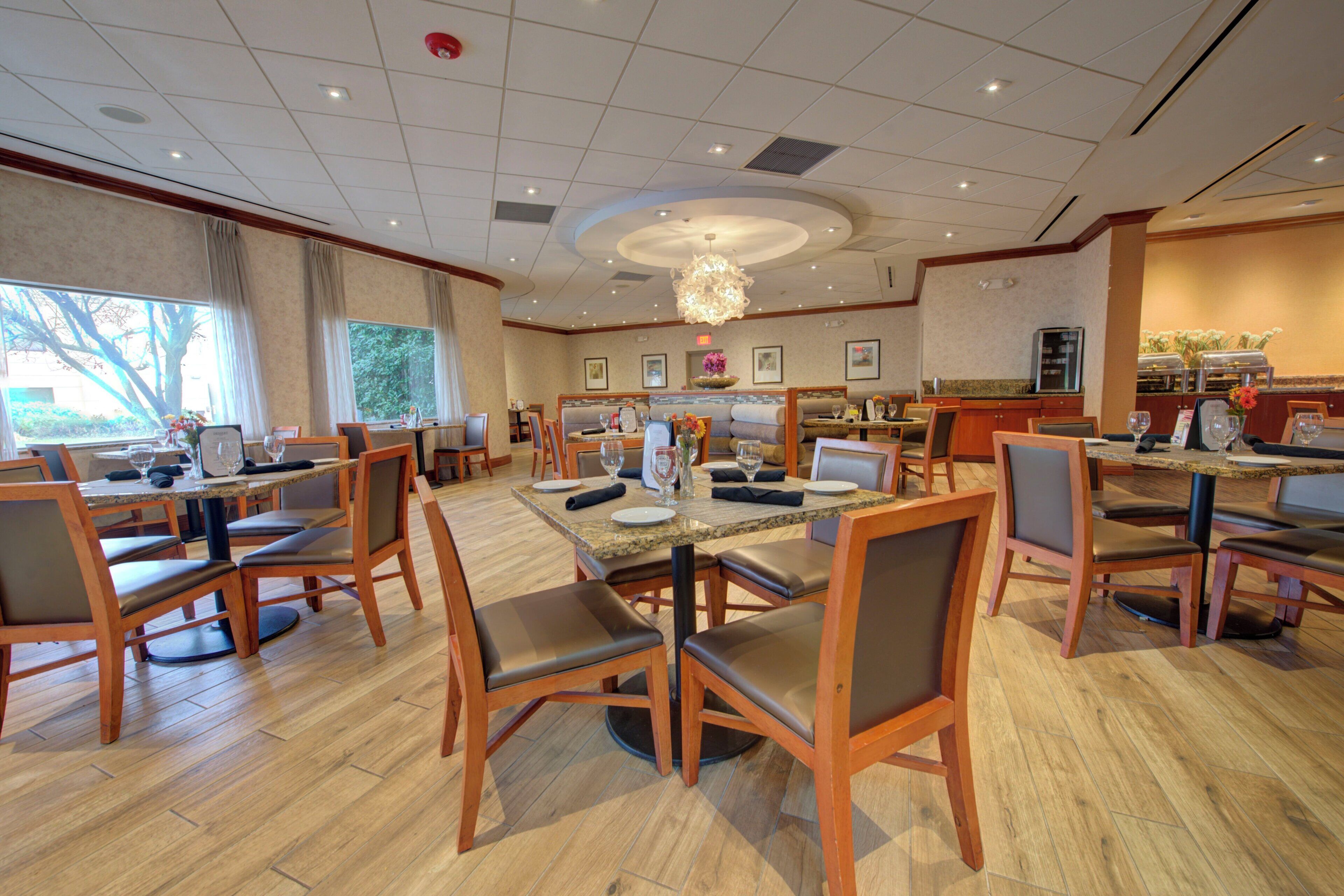 Een restaurantgedeelte in een hotel, met meerdere tafels en stoelen, een groot raam met gordijnen, en een keuken op de achtergrond.