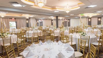 A banquet hall with round tables, gold chairs, and chandeliers.