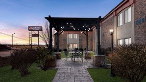 A building with a brick facade, a covered patio area with outdoor seating, and a sign indicating 'Suburban Studios'. - Suburban Studios Kansas City Airport North (Platte City)