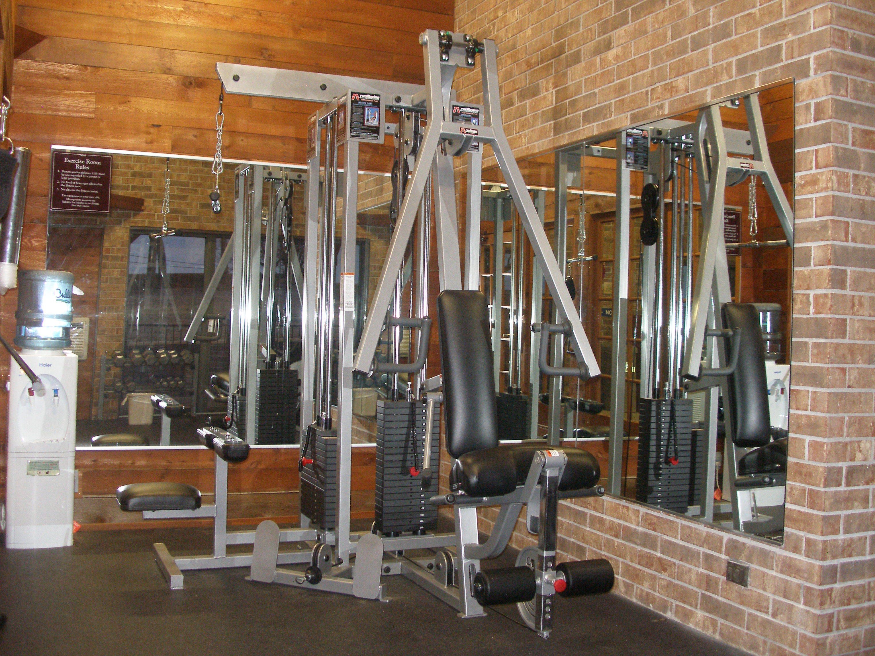 Un gimnasio con diferentes máquinas de ejercicio, un dispensador de agua y un espejo que refleja el equipo.