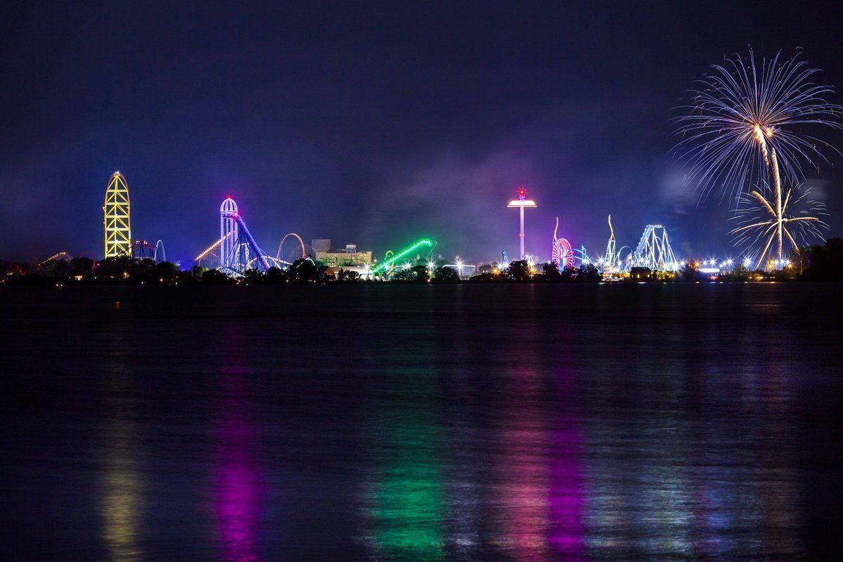 A nighttime view of a waterfront with illuminated amusement park rides and fireworks.