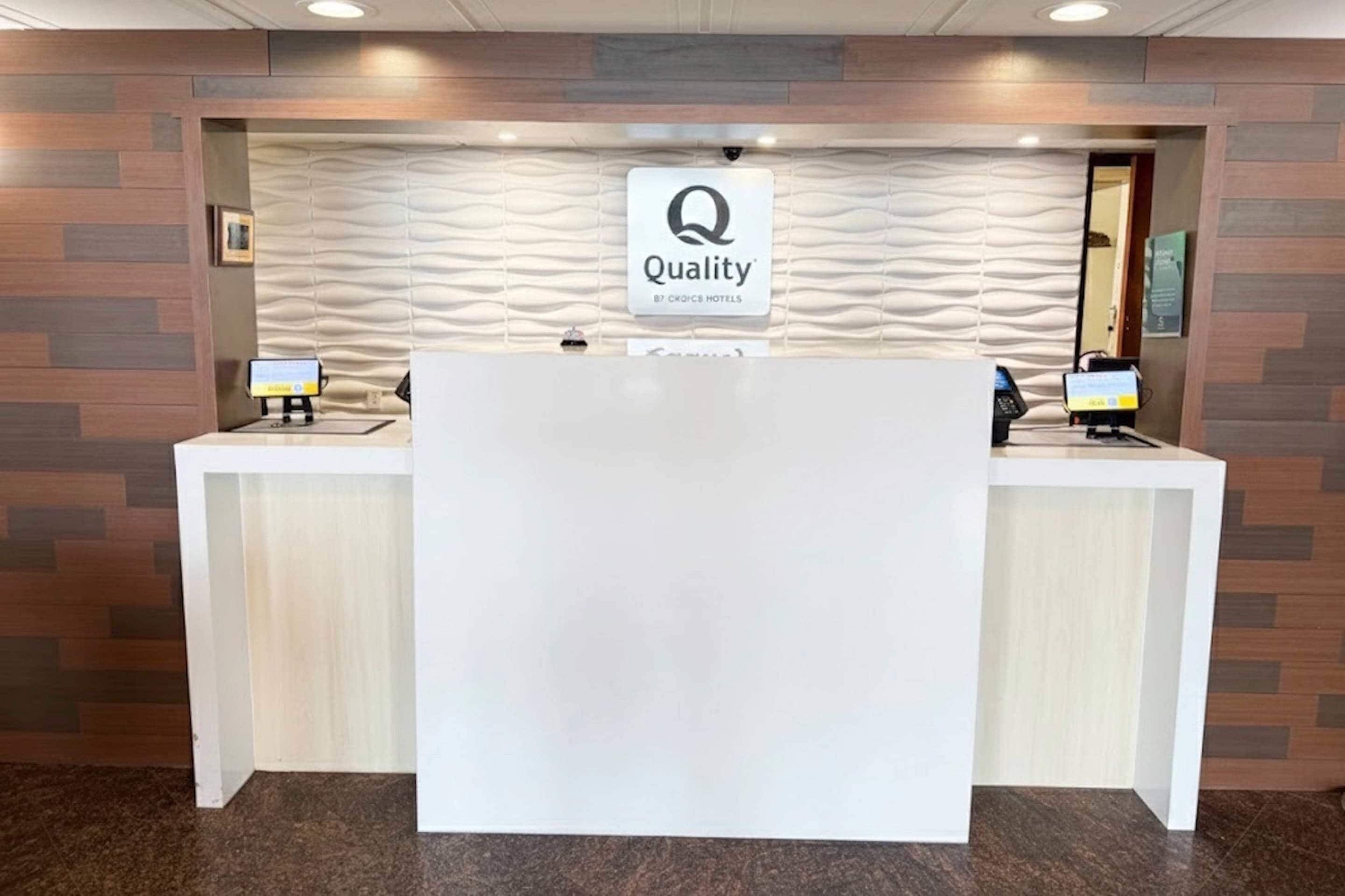 A hotel reception desk with a 'Quality by Choice Hotels' sign, two computer monitors, and a textured wall behind.