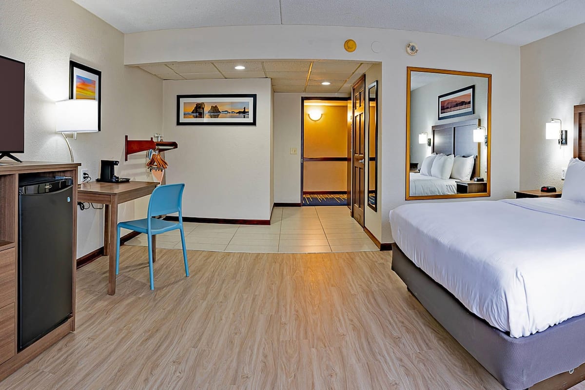 A hotel room with a bed, desk, chair, refrigerator, and a large mirror.