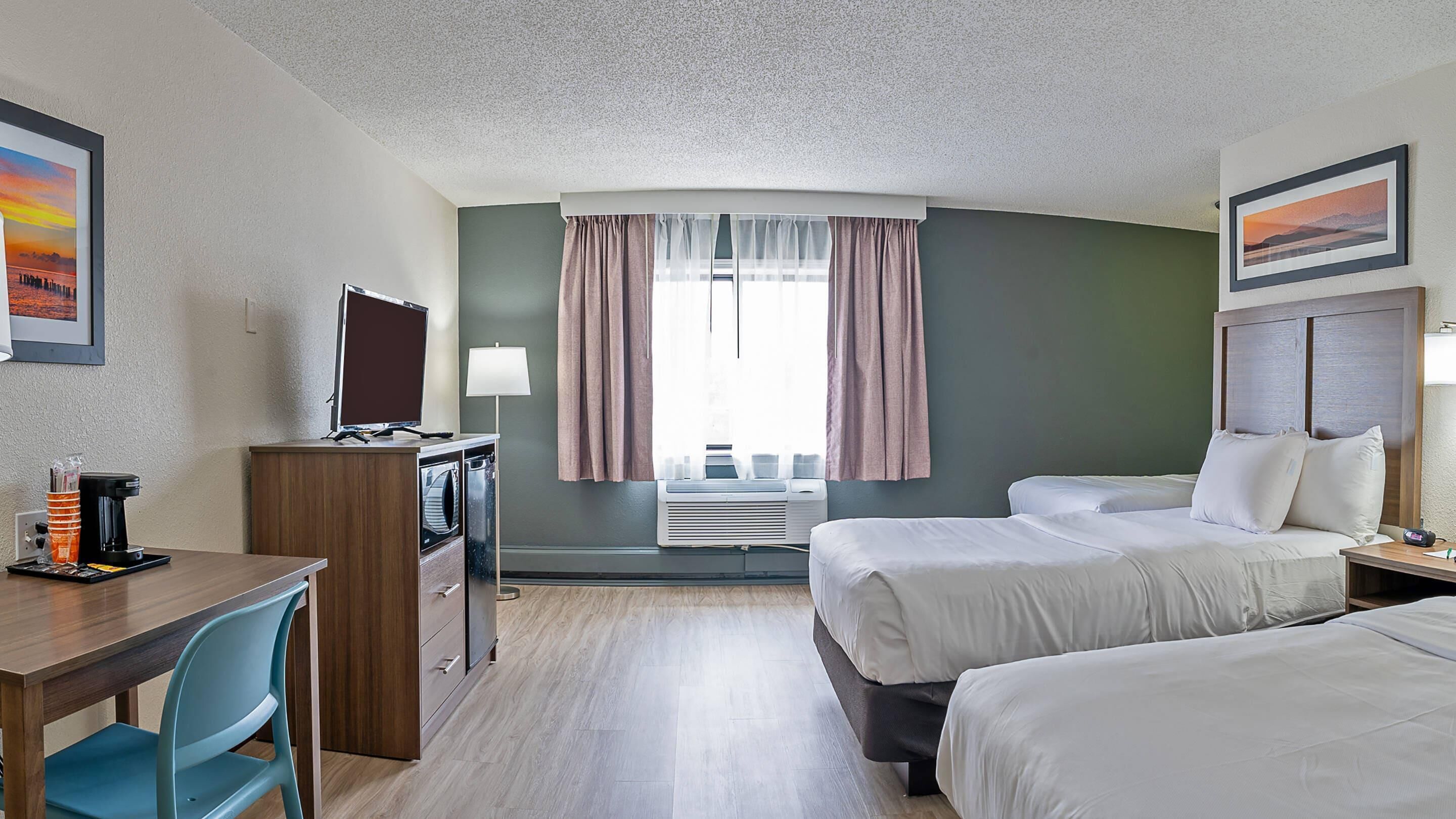 A hotel room with a desk, two beds, a TV, a coffee maker, and a window with curtains.